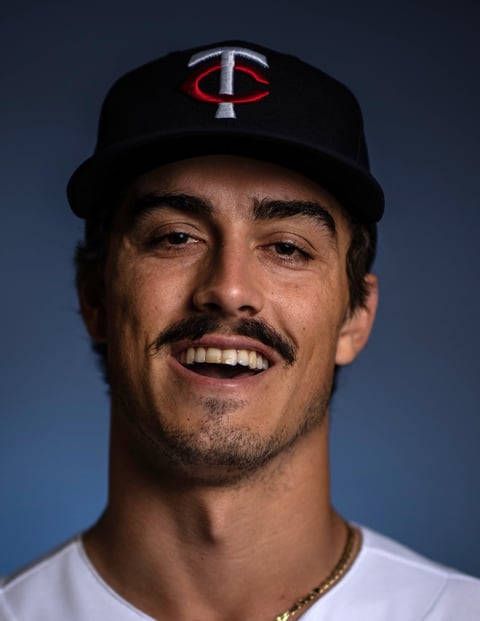 Man smiling at camera wearing black Minnesota Twins baseball cap and white shirt against blue background