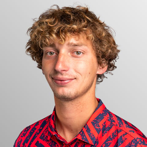 Curly-haired man with a light beard wearing a red and blue patterned shirt against a neutral background