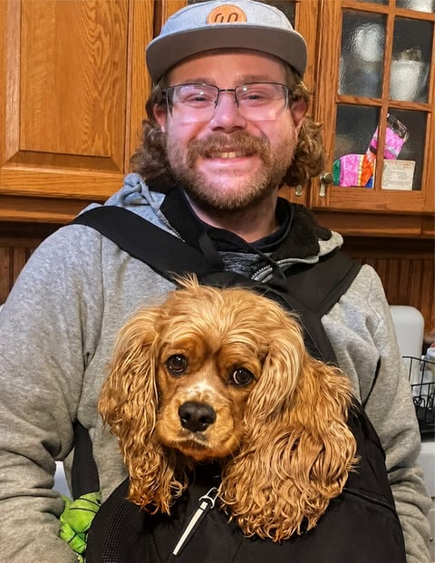 Man wearing cap and glasses holding golden-brown cocker spaniel in kitchen
