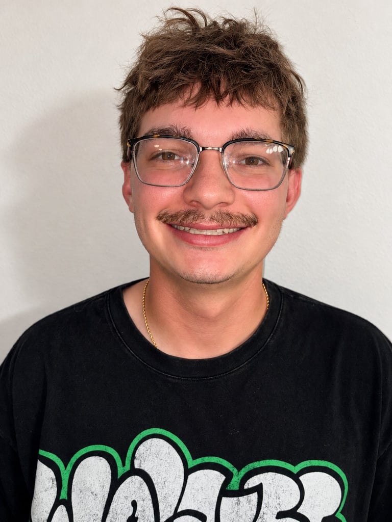 Young man with brown hair and glasses smiling at camera, wearing black t-shirt with green graphic design