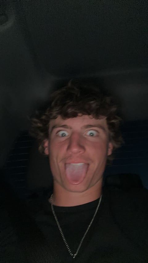 Man with dark curly hair sticking out tongue playfully while making exaggerated facial expression, wearing black shirt and silver chain necklace against dark background